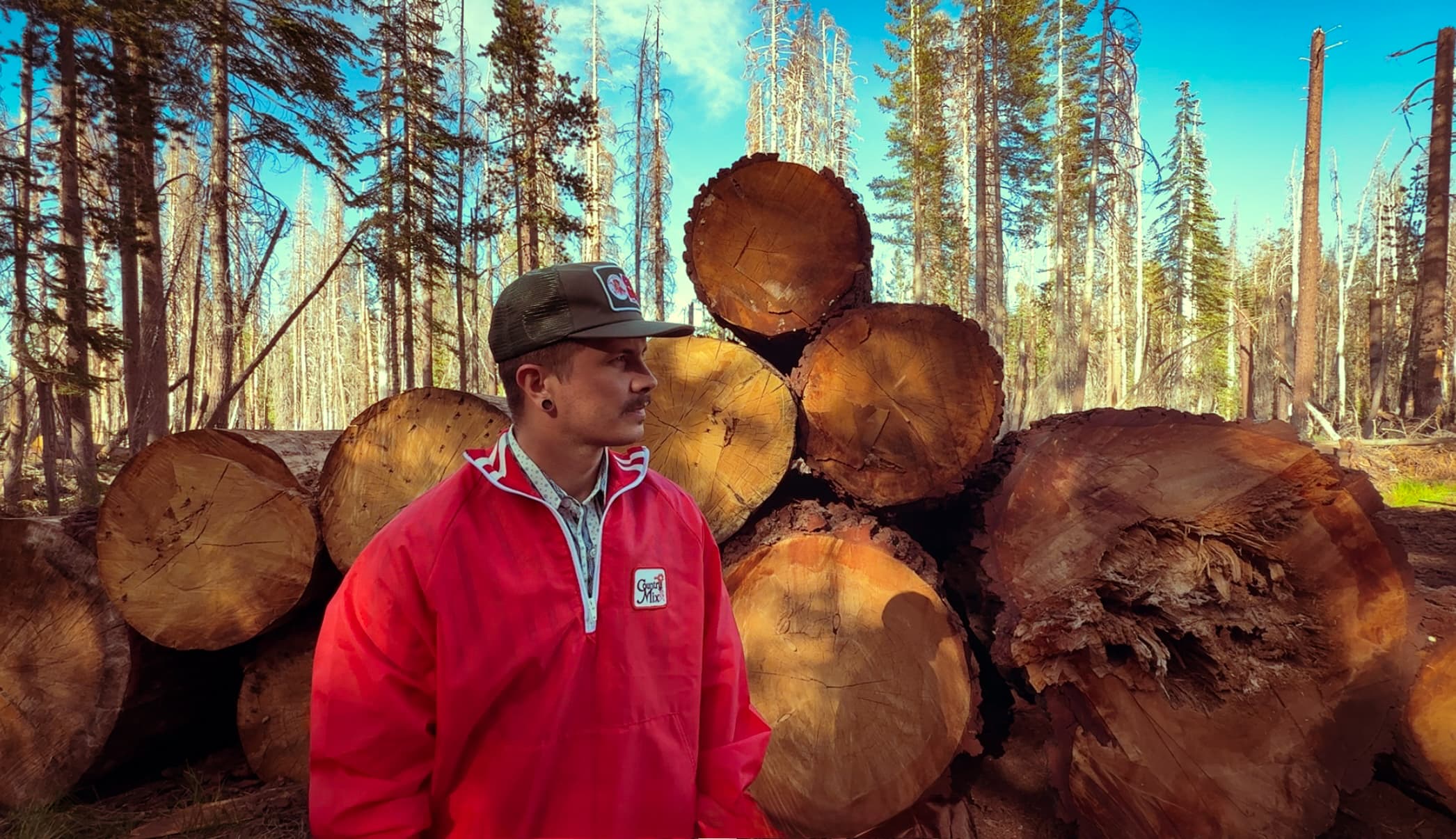 Bridger in a red jacket among a stack of cut logs in a burned forest